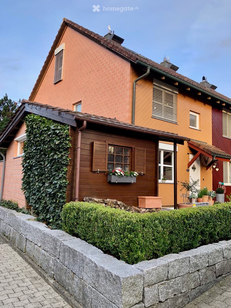 3 story house, brick walls, chimneys, brown roof, windows with shutters, outdoor plants, wall made of grey stones