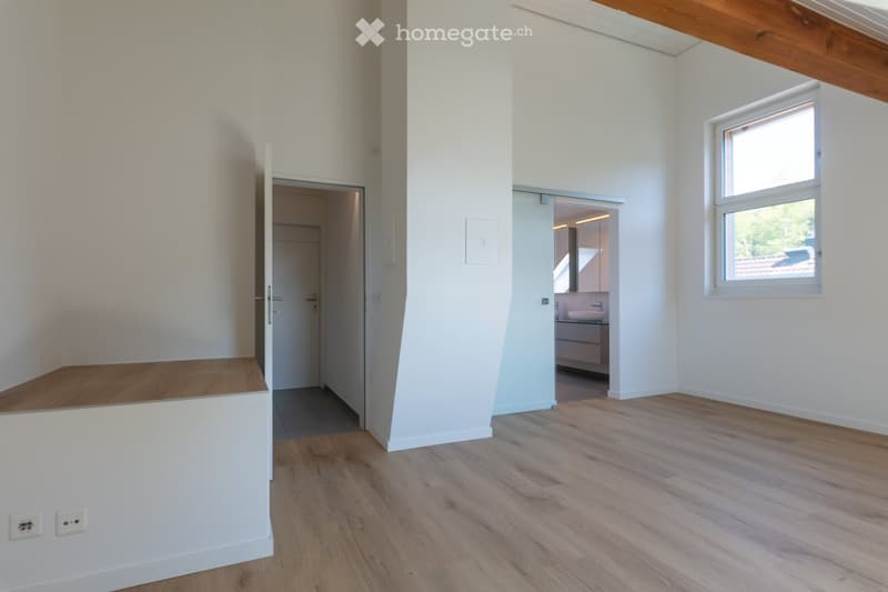 Empty room with wooden floor, white walls, open door, window, power outlets, and a slanted wall.