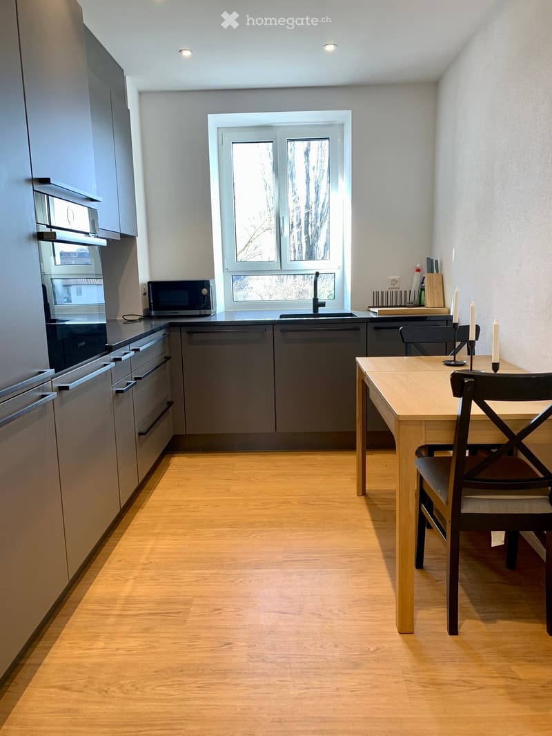 Modern kitchen with oak floors, white walls, grey cabinets, modern appliances, dining table, two chairs, and a sink with a faucet.