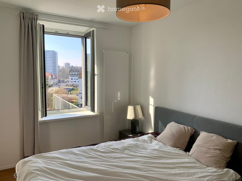 white walls, wooden floor, large window, open window, bed with white sheets, two pillows, bedside lamp