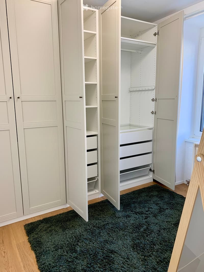 Spacious room with a large white wardrobe with numerous shelves and drawers, wooden floor, and a green carpet.