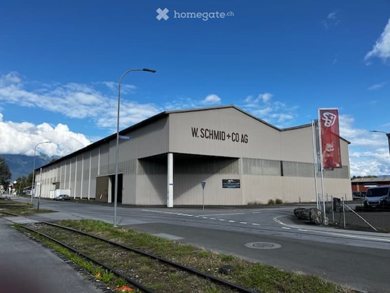 Large industrial building with a sign reading 'W. SCHMID + CO AG', located on a street with a railway track, street lights, and a banner