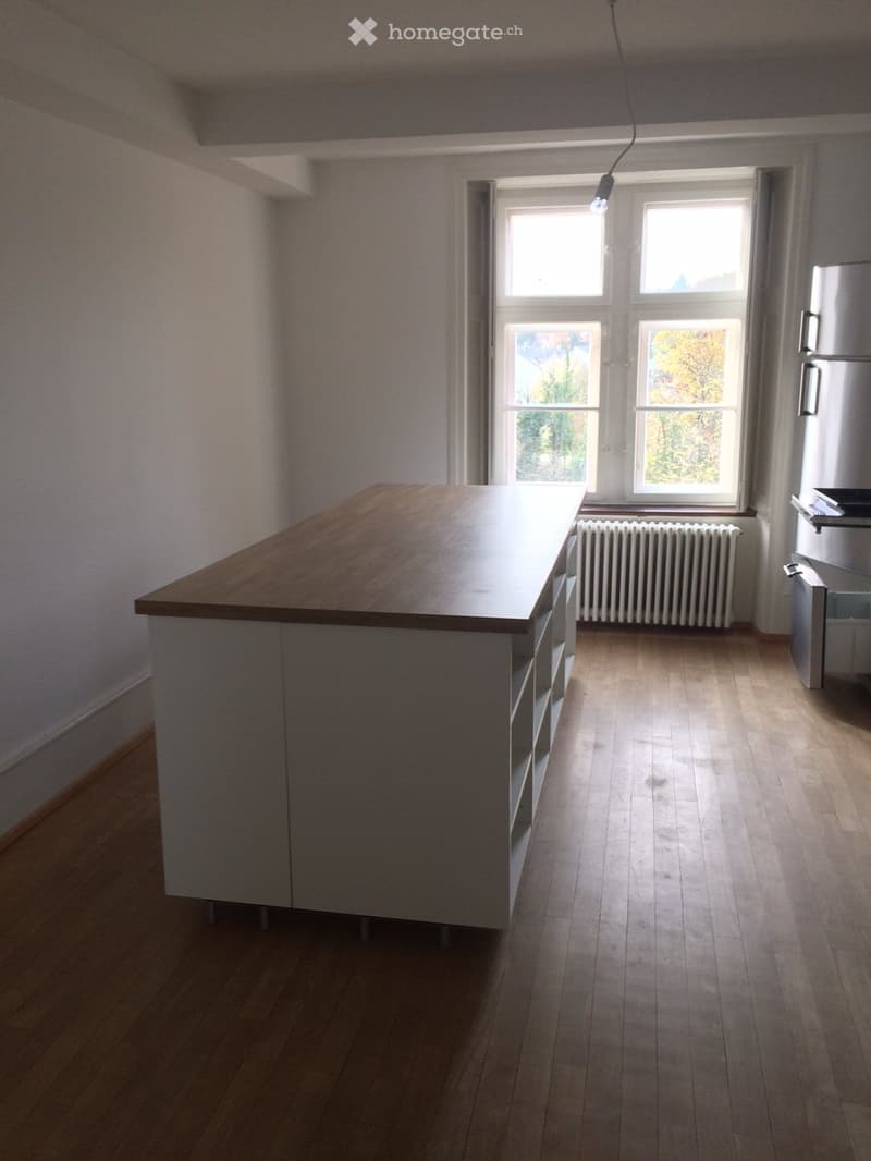 empty kitchen with wooden floor, white walls, large island with shelves, white cabinets, large window, white radiator