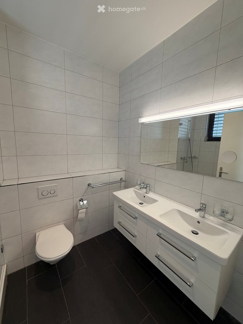 modern bathroom with white tiles, black flooring, large mirror, two sinks, toilet, silver faucets, and vanity drawers