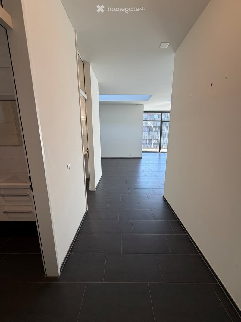 an empty hallway with white walls, black tiled floor, and an open glass door leading to a balcony with a view of the outside