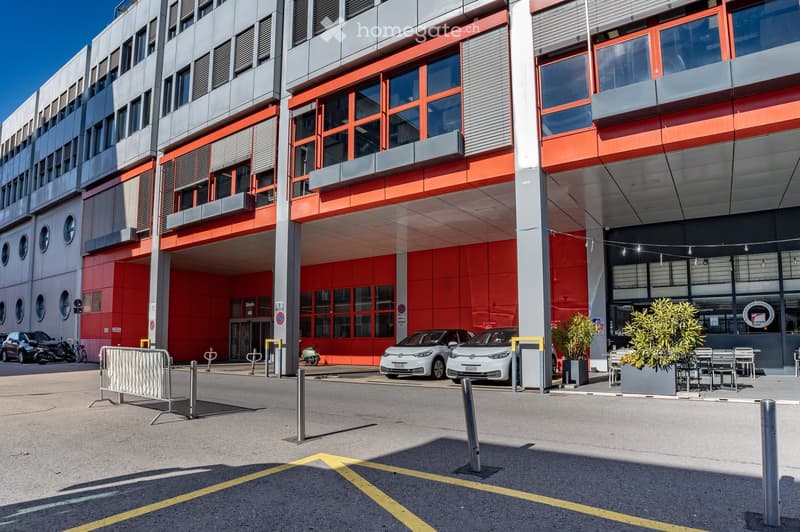 Modern multi-story office building with red accents, multiple windows, designated parking area, outdoor seating, and bollards.