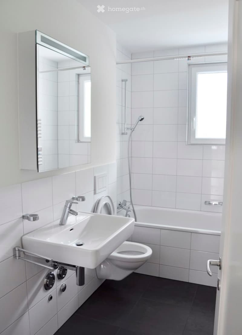 A clean and white modern bathroom with black tile flooring, including a sink, mirror, toilet, bathtub, and shower.