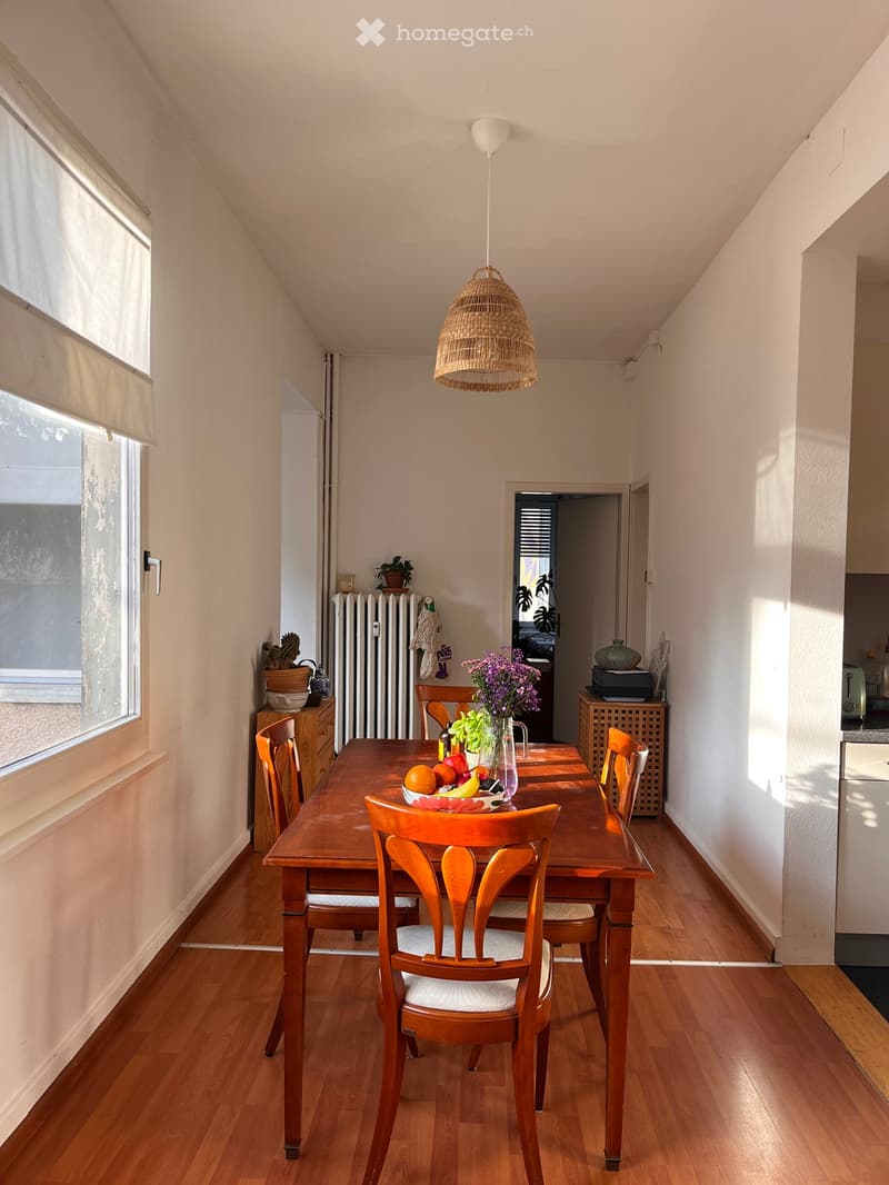 Dining room with wooden table and chairs, white walls, wooden floor, natural light from window, hanging lamp, and various decorative items