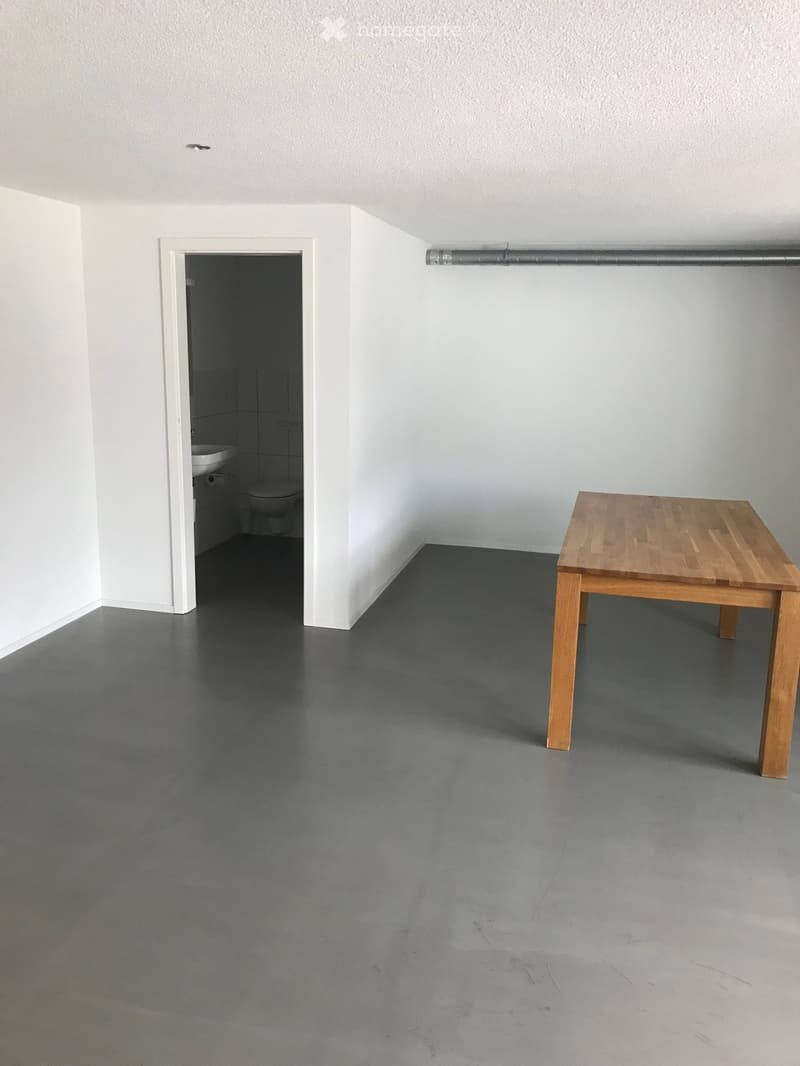 Empty room with white walls, gray floor, wooden table, and a small bathroom with a toilet and sink.