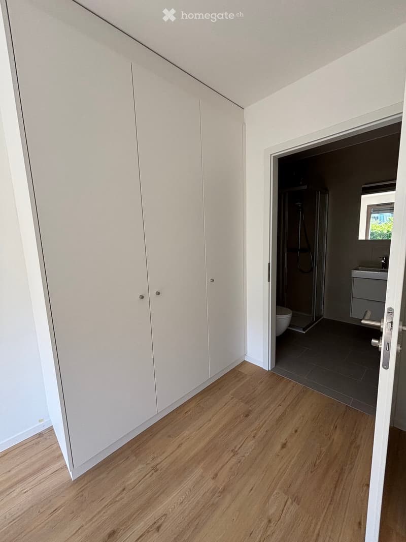A clean, modern hallway with light wooden flooring and white built-in wardrobes. An open doorway leads to a modern bathroom with a shower, sink, and toilet.