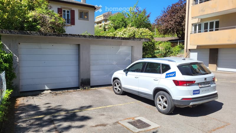 Two white garage doors, a white car parked in front, a building in the background