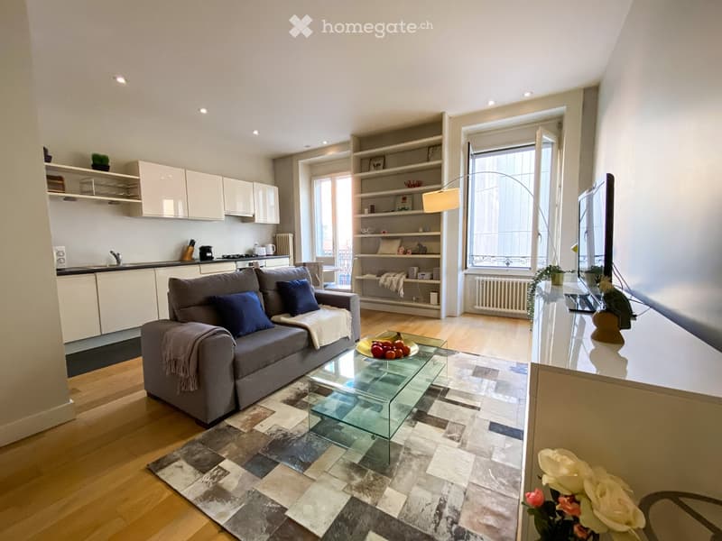 modern living room with gray couch, glass table, wooden floor, white kitchen, large window