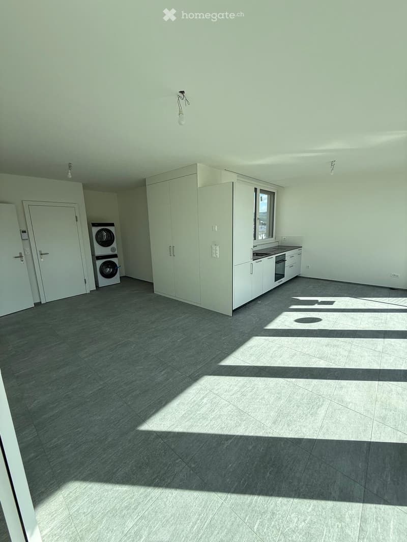 empty room, white walls, gray floor, white cabinets, stacked washing machines, kitchen area with oven