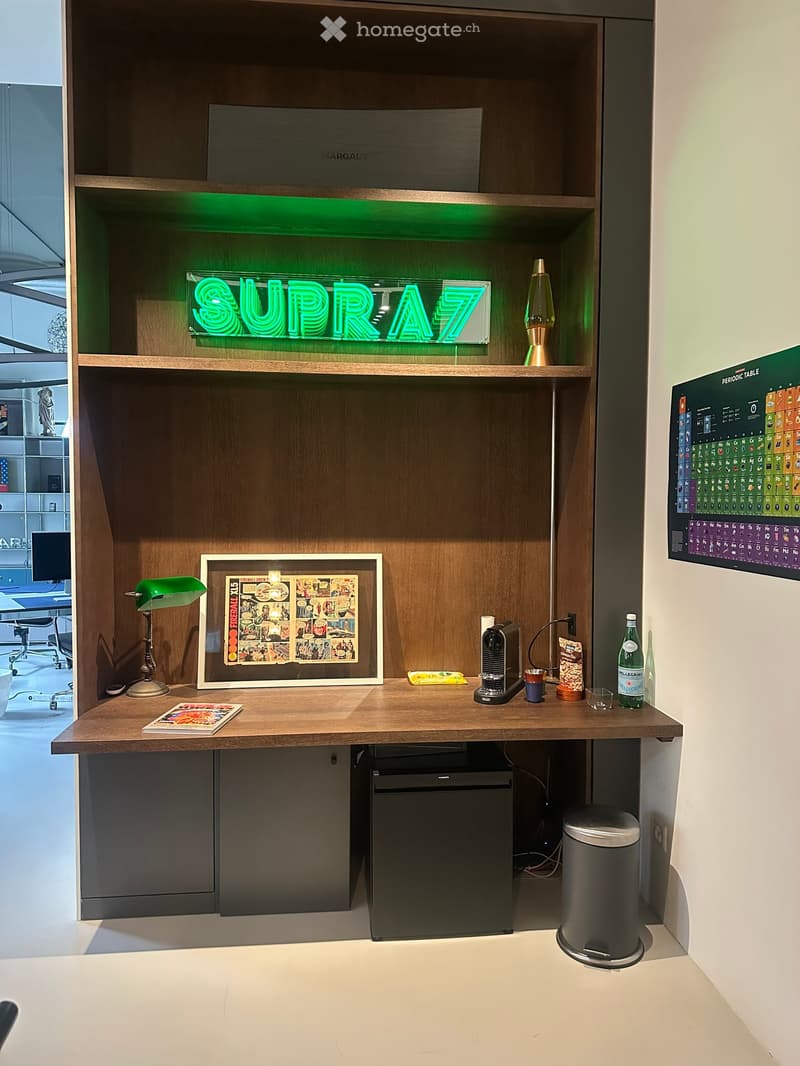A wooden desk with a green neon sign reading 'SUPRA7', a green desk lamp, a framed comic book, a coffee machine, a bottle of water, a trash can, and a mini fridge.