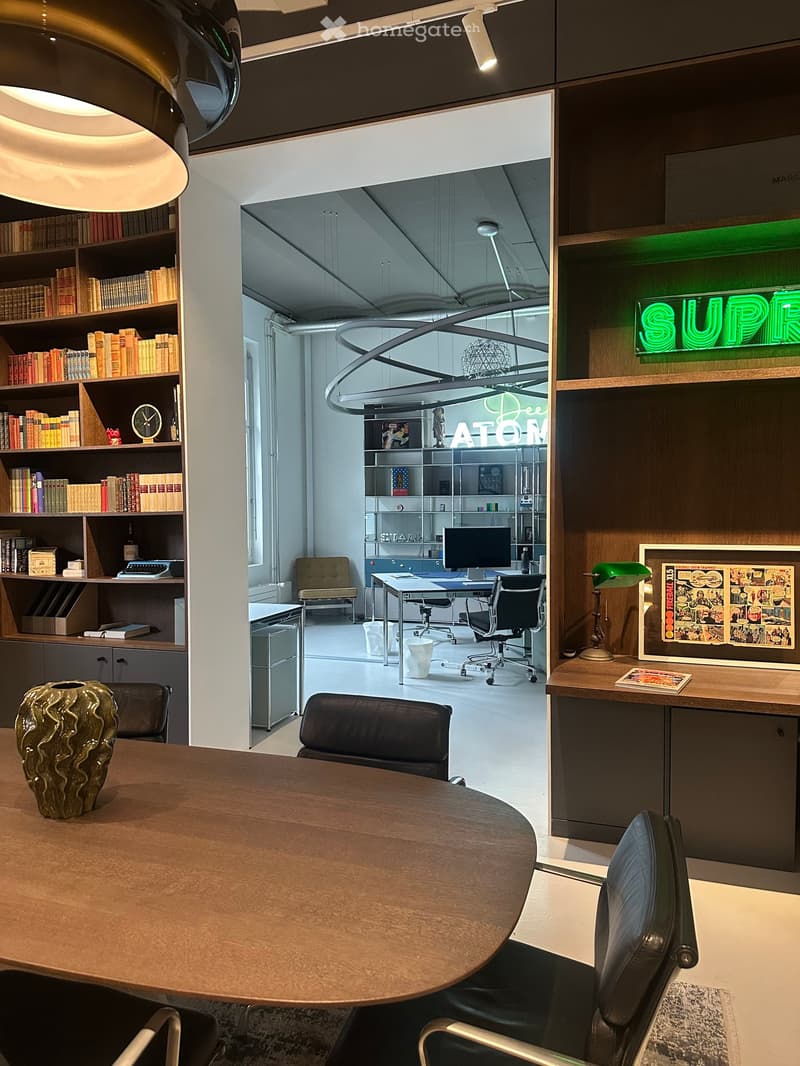 Dining room with a large wooden table, black leather chairs, a large bookshelf filled with books, a green lamp, a large vase, and a framed comic book on the wall.