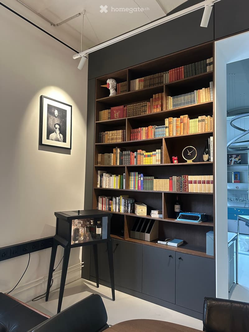 A room with a large bookshelf, a black table with a glass display, a framed picture on the wall, and a black leather chair.