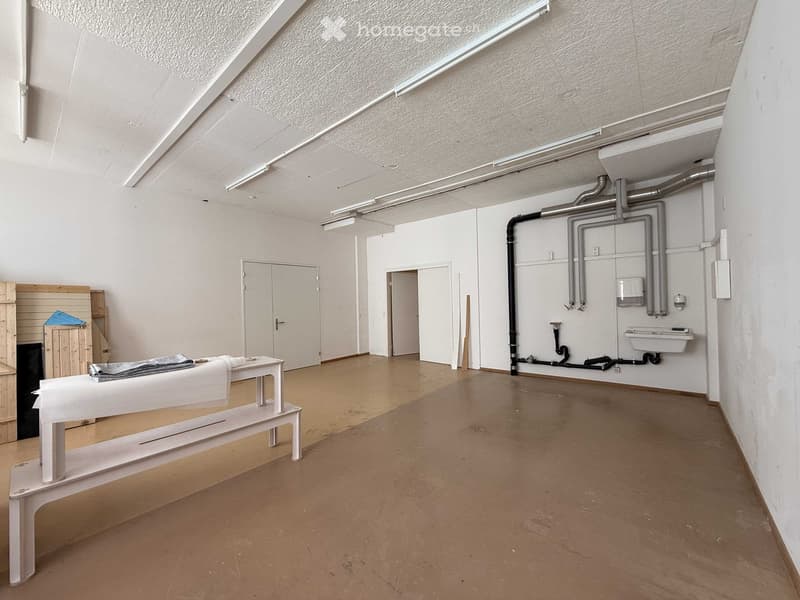 Empty room with white walls, ceiling, and floor. There is a white bench and a wooden storage box in the left corner. The room has a door and a window on the left side. The right side has a sink and pipes on the wall.