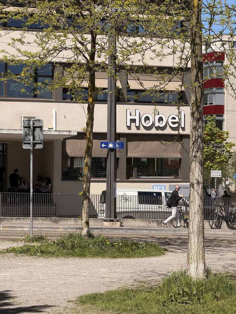 A modern building with a sign reading 'Hobel', people sitting outside, and a man walking with a bicycle.