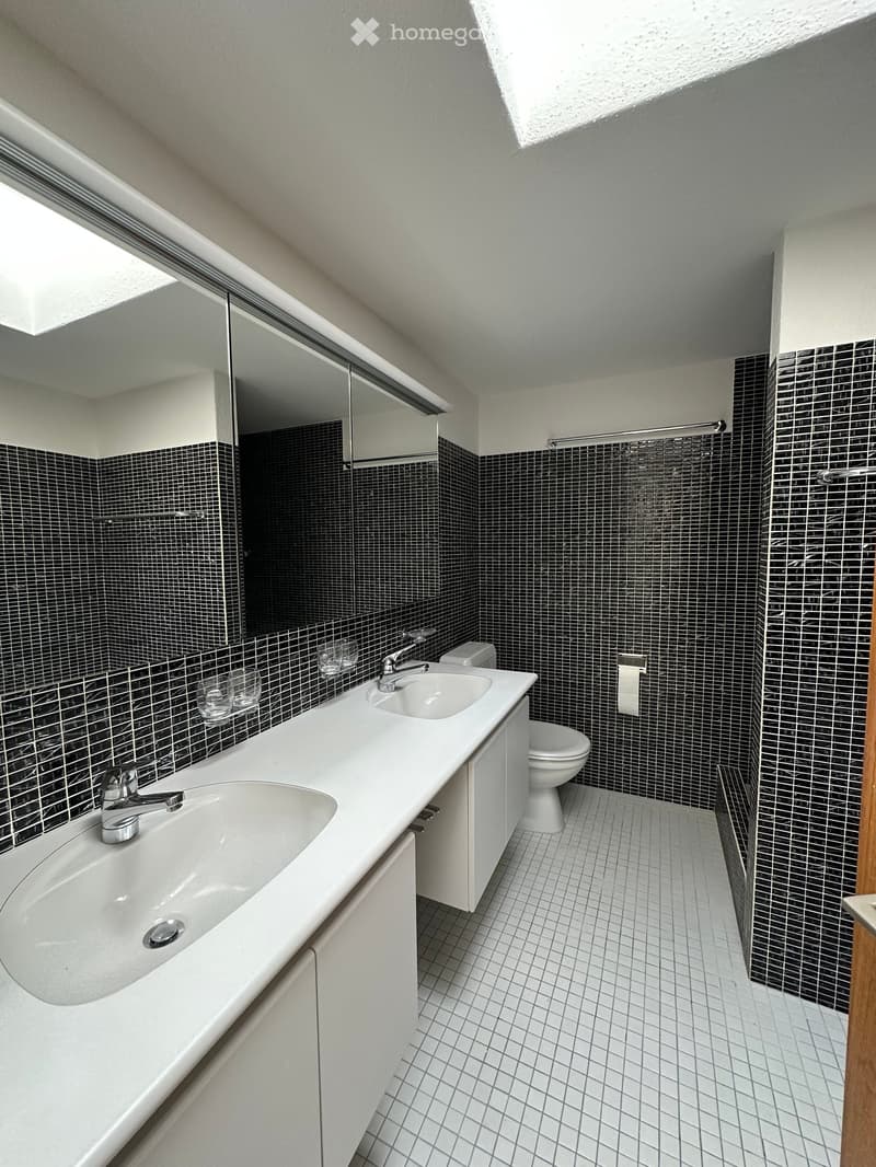 Bathroom with two sinks, a toilet, and a shower. The walls are tiled in black and white, and the floor is tiled in white. There is a mirror above the sinks.