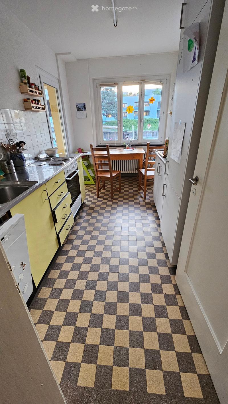 A kitchen with yellow cabinets, a sink, stove, oven, and a wooden table with chairs. The floor is checkered, and there are windows with a view of trees and a building.