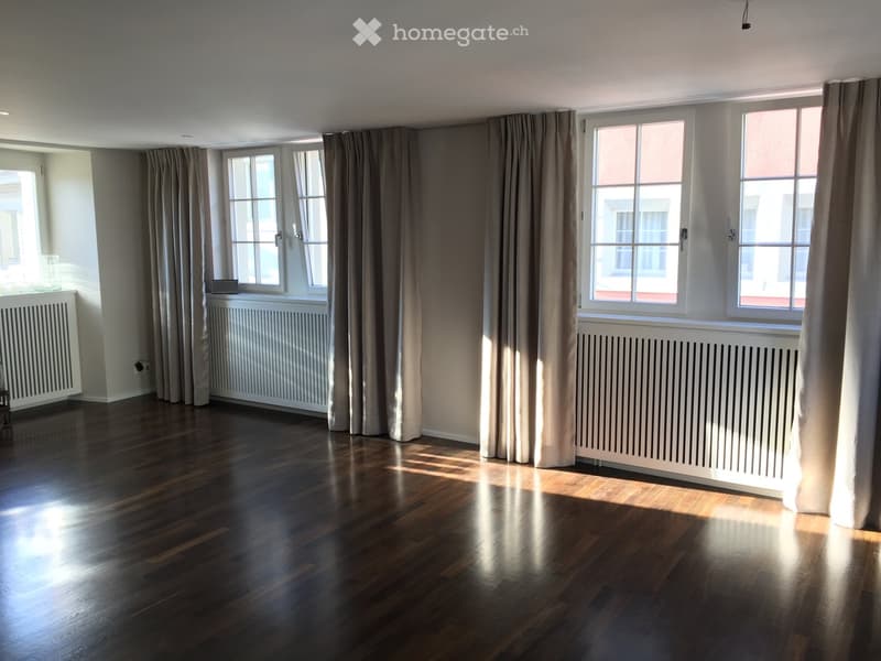 Empty room with hardwood floors, white walls, and large windows covered by curtains. Three white radiators are installed on the walls.