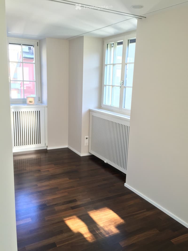 Empty room with white walls, wooden floor, and two windows with white frames. There are white radiators under the windows.