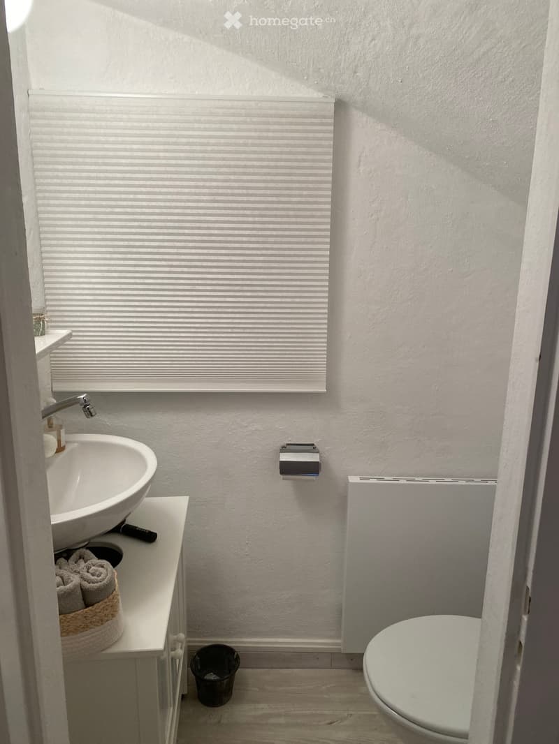 A small bathroom with a sink, toilet, and a towel basket. The walls and floor are white, and there is a window with blinds.
