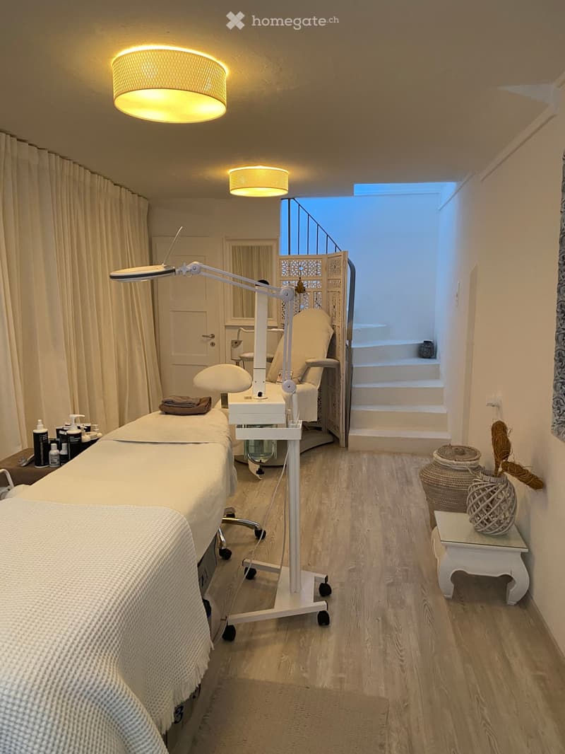 A room with massage tables, lamps, and a chair. The room has a wooden floor and white walls. There is a staircase leading to the upper floor.