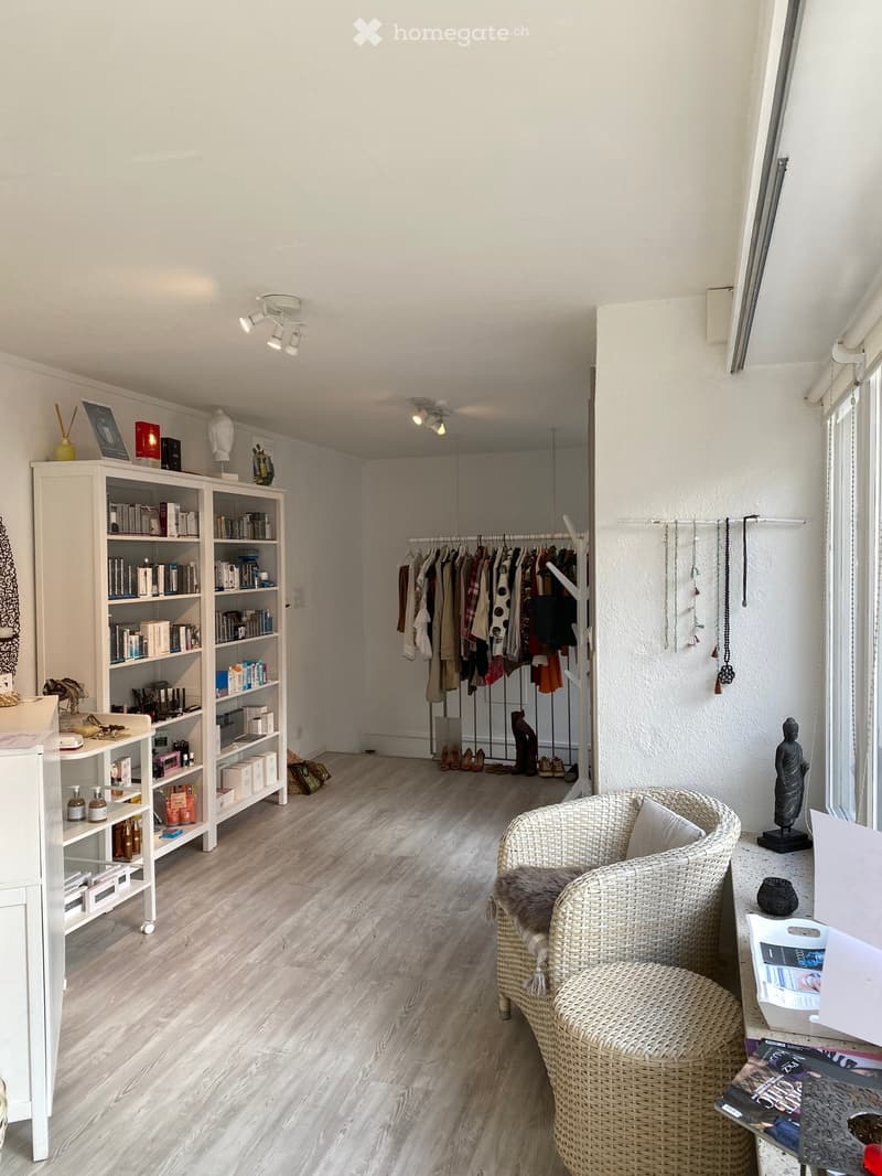 A room with a white bookshelf, a chair, a table, a sculpture, a clothes rack, shoes, necklaces, a candle, books, bottles, a basket, magazines, and a lamp.