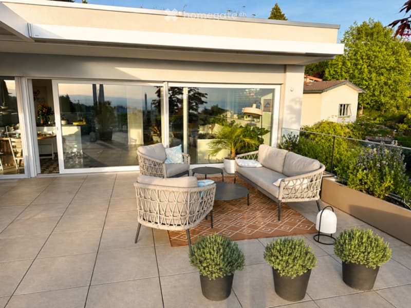 Outdoor terrace with a seating area, featuring woven chairs, a round table, a rug, and potted plants.