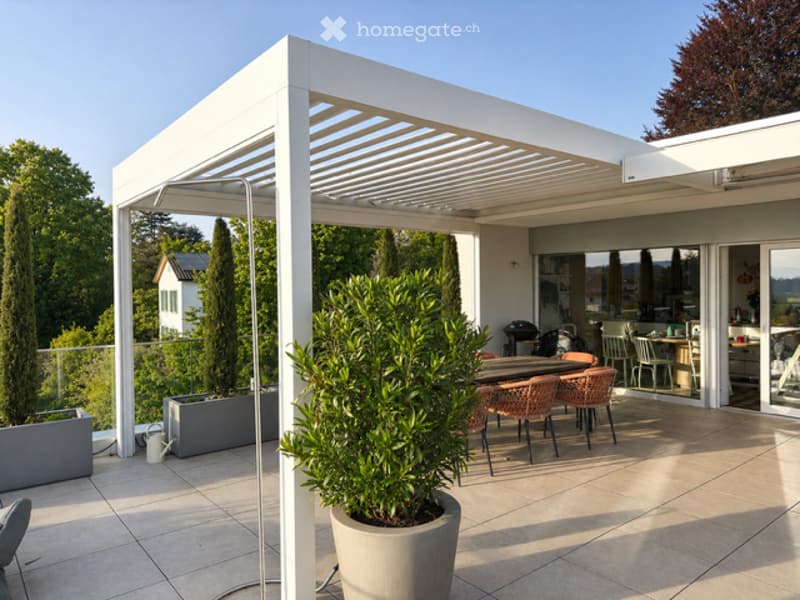 Outdoor dining area with a white pergola, tiled floor, and a large potted plant. There are chairs and a table set up for dining.