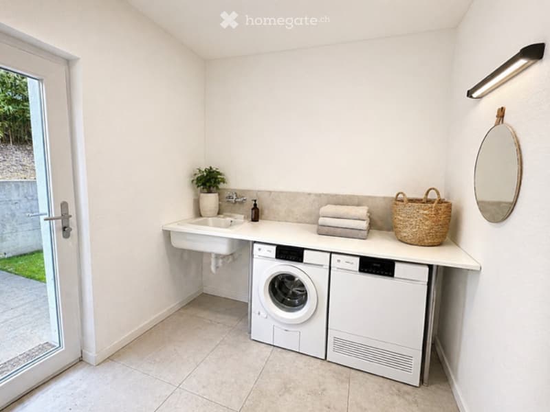 A small laundry room with a washing machine, dryer, sink, and a basket. The room has white walls and tiled flooring.