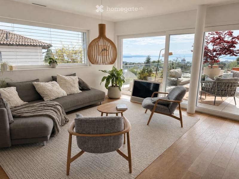 Spacious living room with a gray sofa, wooden chairs, coffee table, and a rug. Large windows provide a view of the outdoors. Potted plants and a lamp add to the decor.