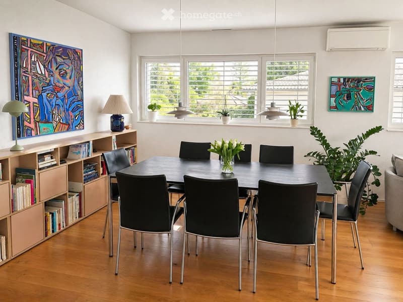 Dining area with a black table and chairs, wooden floor, white walls, and large windows with blinds. A bookshelf filled with books and two paintings on the wall. A plant and a vase with flowers on the table.