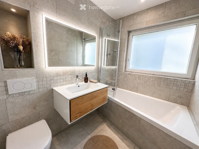 Modern bathroom with a white bathtub, a white sink, a large mirror, and a shower area. The floor is tiled, and there is a toilet and a rug in the room.