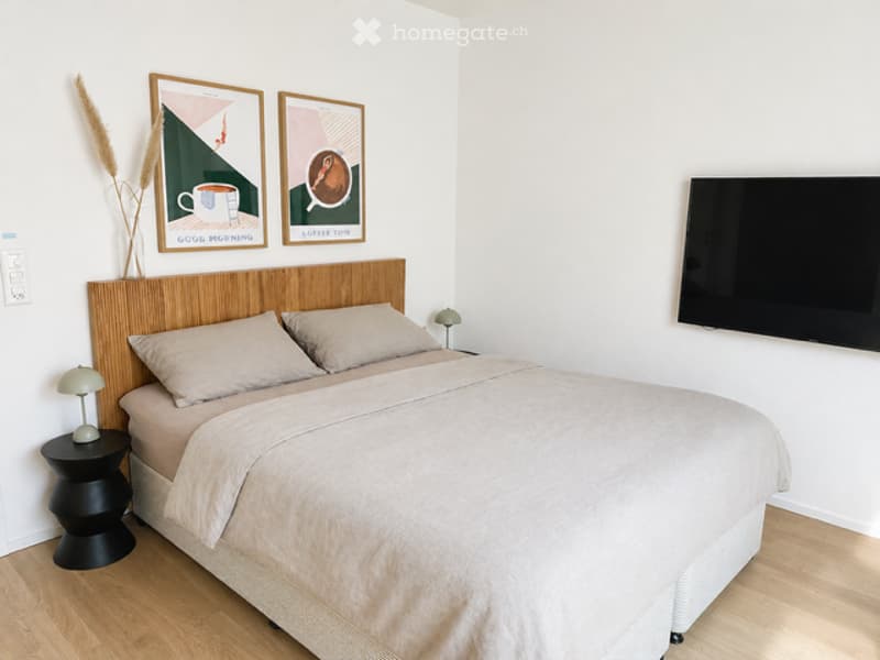 A bedroom with a bed, pillows, nightstand, lamp, and a television mounted on the wall.