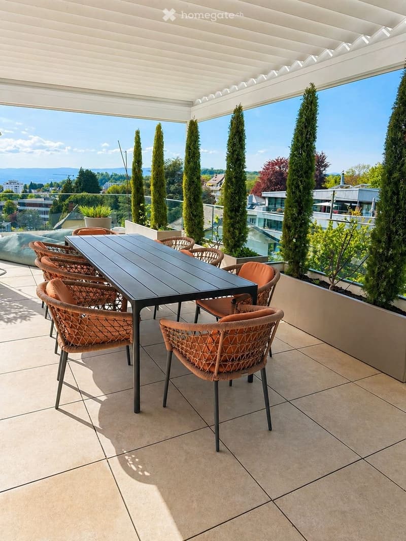 Outdoor dining area with a black table and brown chairs, surrounded by tall cypress trees and planters. The balcony has a tiled floor and a view of the city.