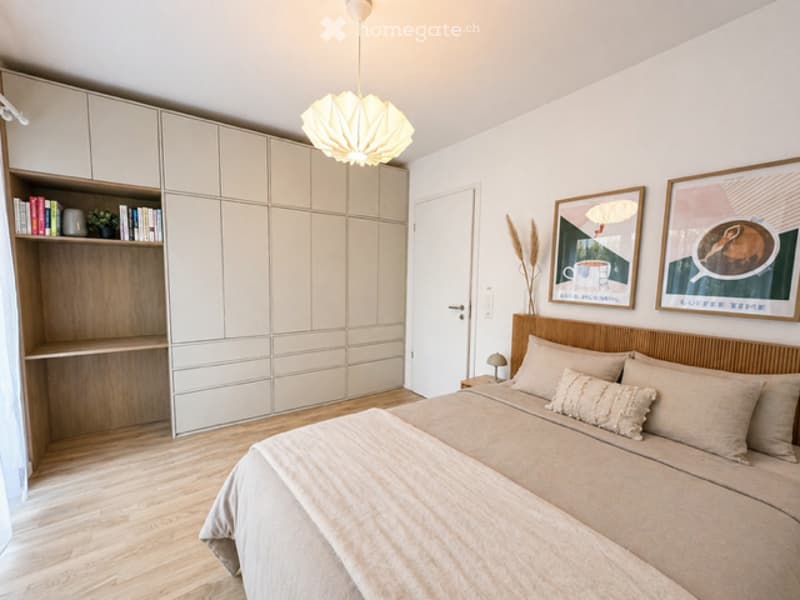 A bedroom with a bed, nightstand, and a wooden headboard. The room has a wooden floor and white walls. There is a bookshelf with books and a plant on it. There are two paintings on the wall, and a lamp is on the nightstand. The room has a door and a cabin
