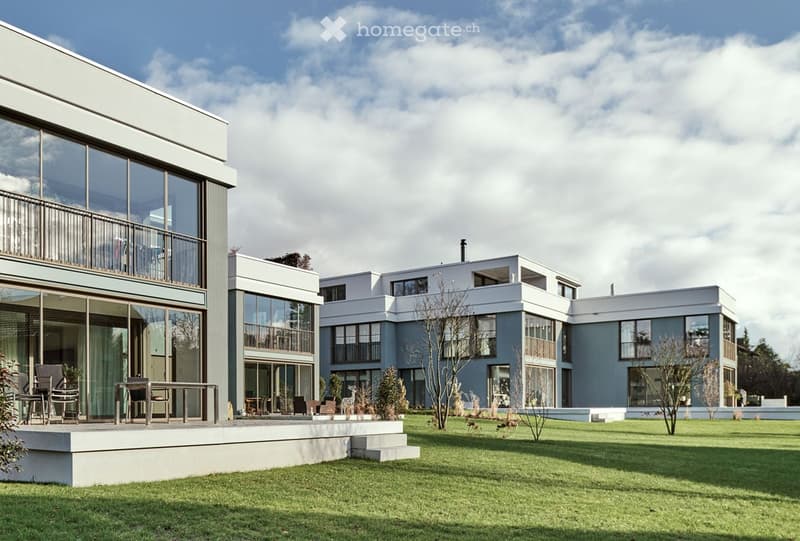 Modern architectural building with large glass windows and balconies, set in a landscaped garden.