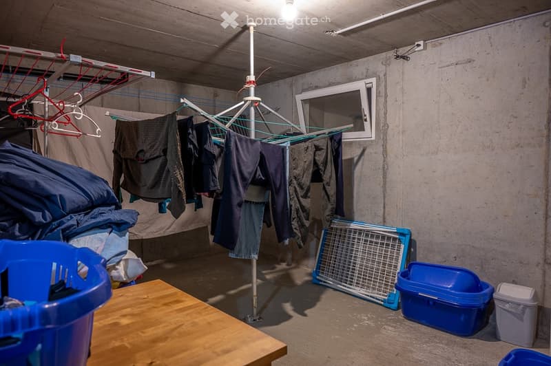 A room with concrete walls and floor, containing a clothes drying rack, a table, and storage bins.