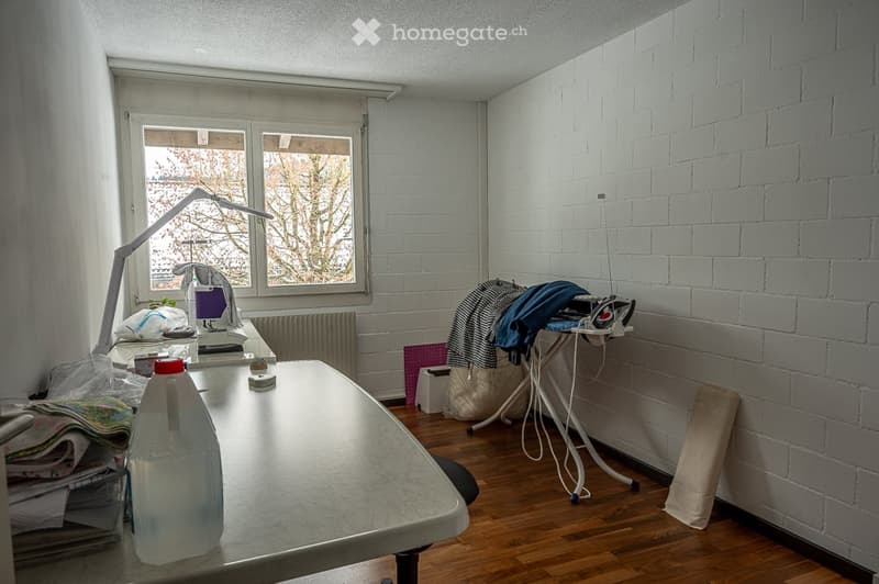 A room with a desk, lamp, and ironing board. There are clothes on the ironing board and a plastic bag on the desk. A bottle, storage box, and basket are also present.