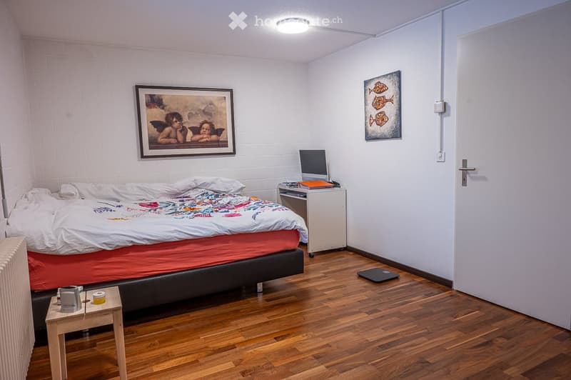 A bedroom with a bed, nightstand, desk, computer monitor, lamp, paintings, radiator, scale, bottle, and cup.