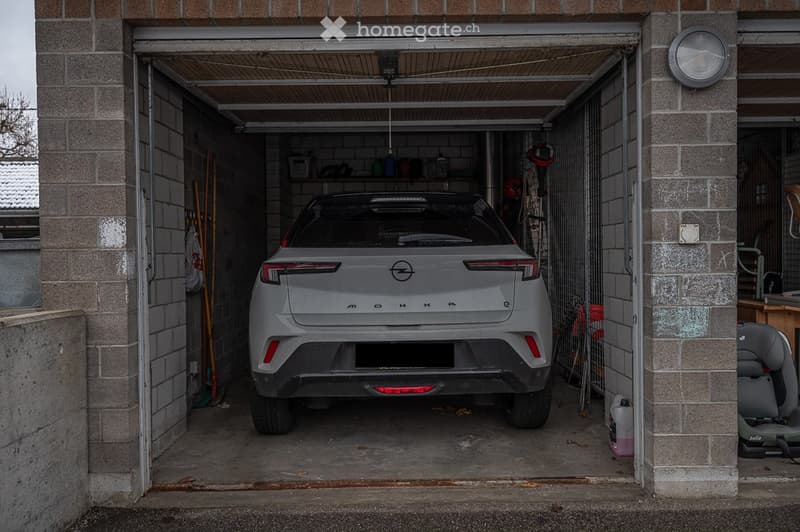 A car parked inside a garage with a brick wall, a shelf with various items, and a ladder leaning against the wall.
