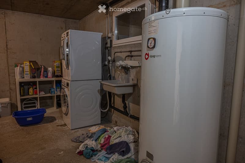 A laundry room with a washing machine, dryer, sink, and water heater. There is a pile of laundry on the floor and a shelf with various items.