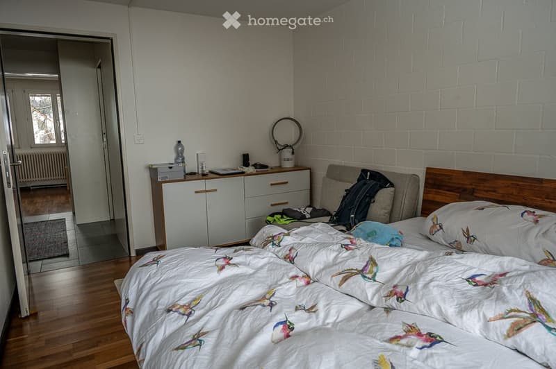Bedroom with a bed, nightstand, cabinet, and chair. The bed has a white comforter with colorful bird designs. The nightstand has a bottle and books. The cabinet has drawers and a mirror on top. The chair has a backpack on it. The room has a wooden floor a