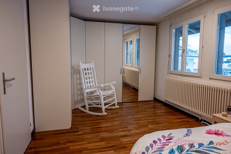 A bedroom with a white rocking chair, a bed, and a white wardrobe. The room has a wooden floor and white walls. There is a radiator on the wall and a window with a view of the outside.