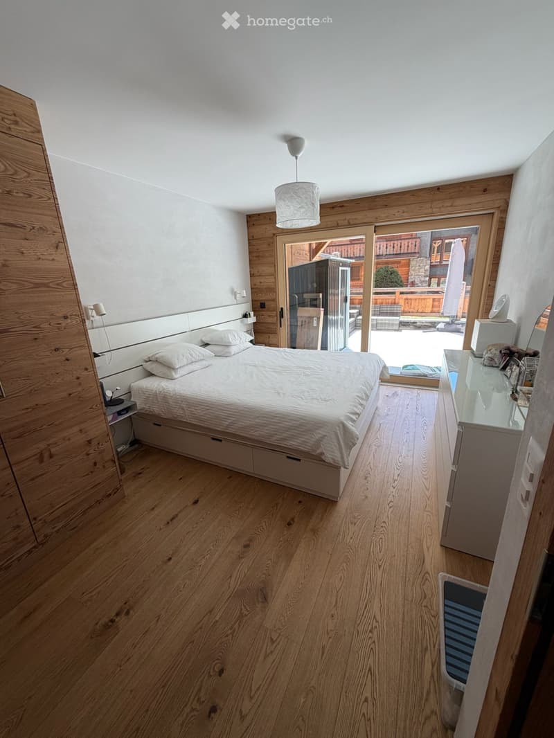 Spacious bedroom with wooden floors, a large bed, and white walls. The bed has a white headboard and is made up with white sheets and pillows. There is a nightstand with a lamp on the left side of the bed. A wooden wardrobe is on the left side of the room