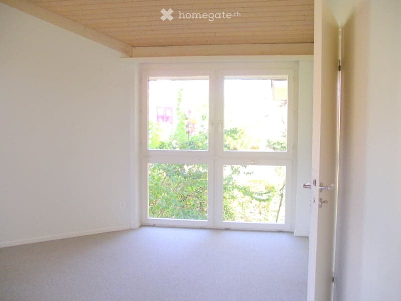 Empty room with white walls, wooden ceiling, and carpeted floor. Large windows with a view of trees outside.