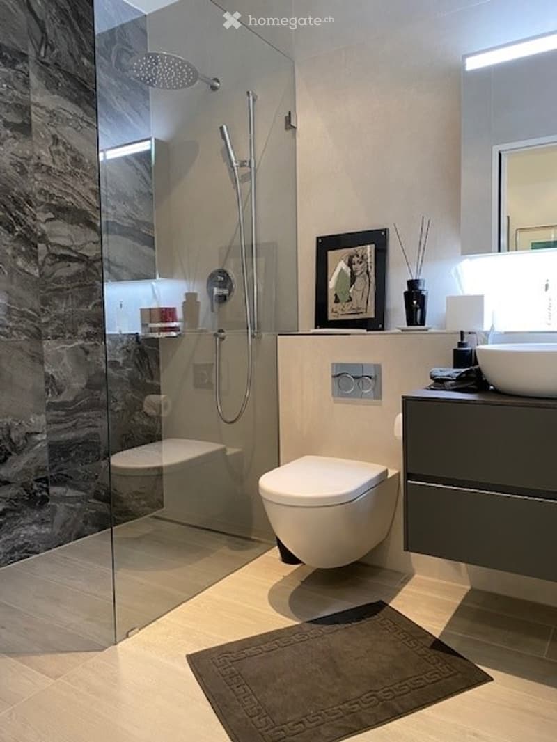 Modern bathroom with a glass shower enclosure, a white toilet, a white sink, and a gray cabinet. The floor is tiled with light-colored tiles, and there is a rug in front of the toilet.
