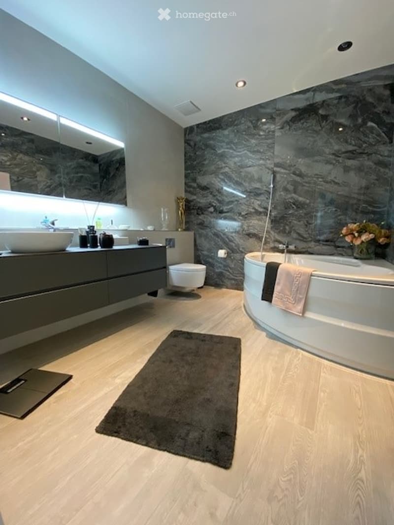 Modern bathroom with marble walls, wooden floor, large mirror, white sink, and bathtub with towels.